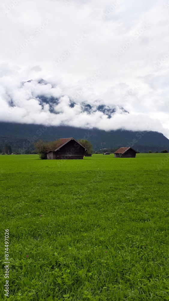 Misty mountain peaks looming over a lush green meadow with traditional wooden huts, creating a serene and picturesque landscape, Garmisch-partenkirchen, Germany Misty mountain peaks looming over a lush green meadow with traditional wooden huts, creating a serene and picturesque landscape, Garmisch-partenkirchen, Germany