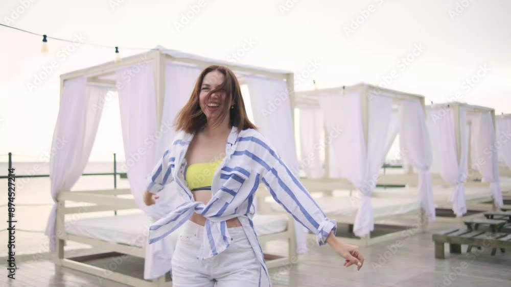 A cheerful, young blonde woman wearing summer striped shirt dances with the rainy wind sunrise at beachside resort A cheerful, young blonde woman wearing summer striped shirt dances with the rainy wind sunrise at beachside resort