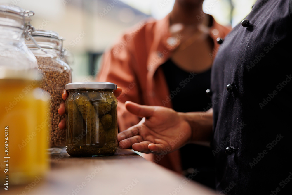 Closeup of african american person with an apron at local market selling fresh eco-friendly produce, advising customer on sustainable food choices. Female seller showing client glass jar of organic Closeup of african american person with an apron at local market selling fresh eco-friendly produce, advising customer on sustainable food choices. Female seller showing client glass jar of organic