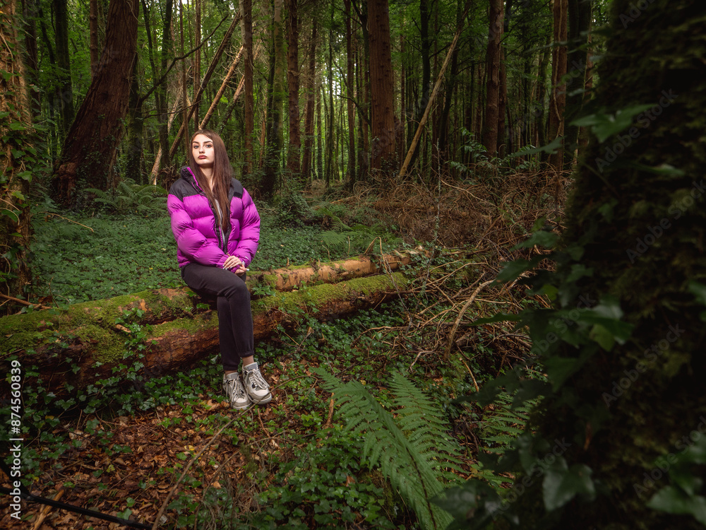 Young teenager girl in a dark forest park sitting on a tree. Travel and tourism concept. Model is with long dark hair and slim body type wearing colorful jacket and black trousers. Young teenager girl in a dark forest park sitting on a tree. Travel and tourism concept. Model is with long dark hair and slim body type wearing colorful jacket and black trousers.