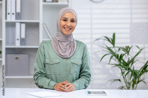 Confident businesswoman wearing hijab sitting at office desk with notepad and tablet