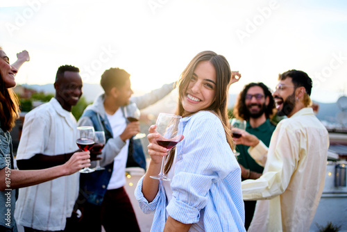Foto Young Caucasian pretty woman poses coquette holding red wine with friends in background dancing at rooftop