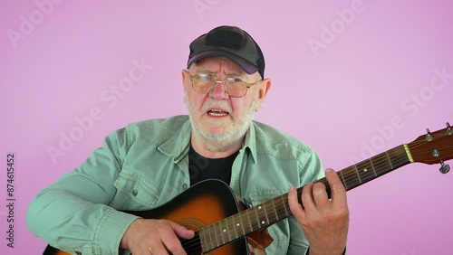 An elderly man in a baseball cap, glasses, plays an acoustic guitar and sings a song. Slow motion. Green background.
