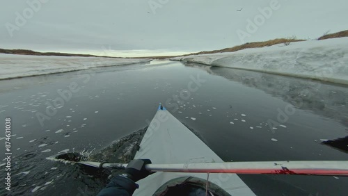 A kayaker paddles through a narrow, icy river, the snow-covered banks towering over them. The cold, clear water reflects the winter sky, creating a serene, otherworldly scene