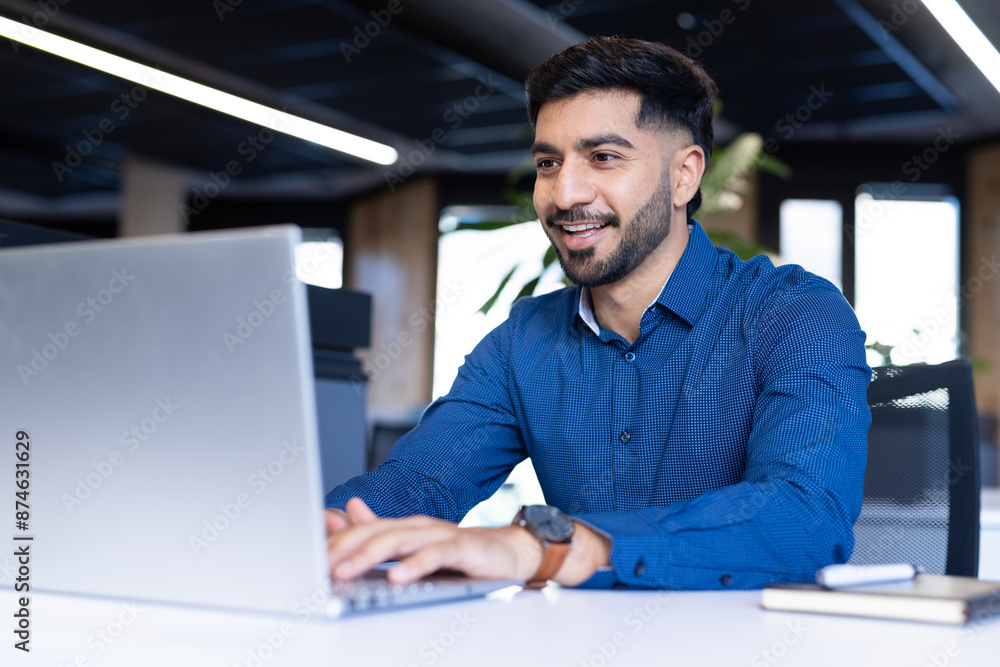 Working on laptop, smiling businessman in blue shirt at modern office desk Working on laptop, smiling businessman in blue shirt at modern office desk