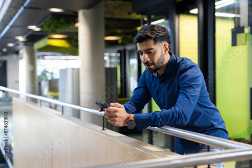 Using smartphone, man leaning on railing in modern office building, copy space
