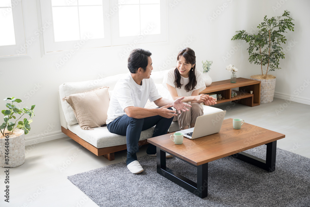 明るい綺麗なリビングでパソコンを見ながら話し合う50代夫婦　不動産や旅行を探すイメージ広角　