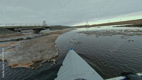 First-person view kayak paddling through icy waters towards a bridge on a cloudy day.