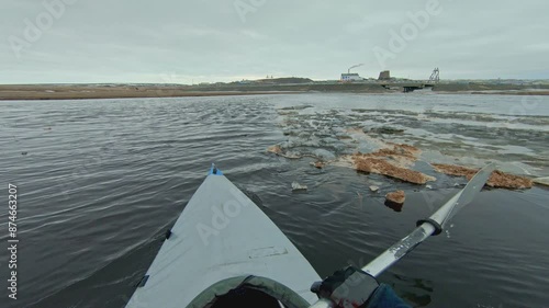Point of view shot from a kayak traveling up a river towards a bridge. The water is dark and there are small chunks of ice floating in it. The shore is brown and barren and the sky is overcast.