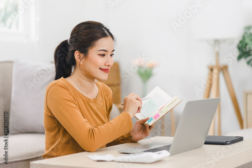 Smiling asian woman working on laptop while sitting in comfortable home with notebook and documents. © kenchiro168