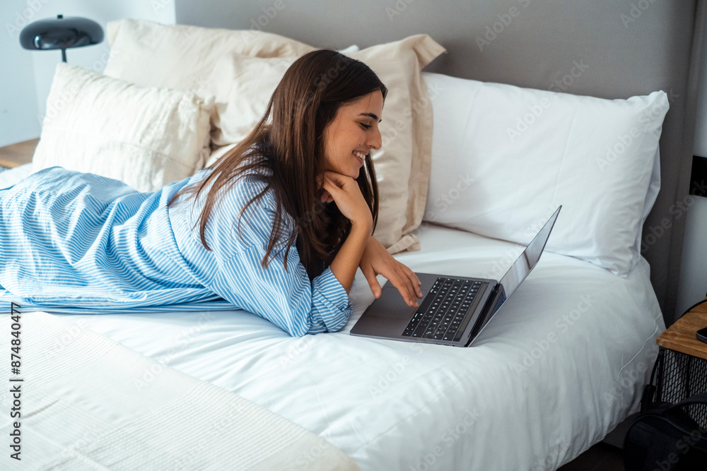 Lazy modern young woman using social media on laptop laying on bed in morning wake up wearing casual clothes and smiling at the display. Working remote with computer and wireless connection in hotel Lazy modern young woman using social media on laptop laying on bed in morning wake up wearing casual clothes and smiling at the display. Working remote with computer and wireless connection in hotel