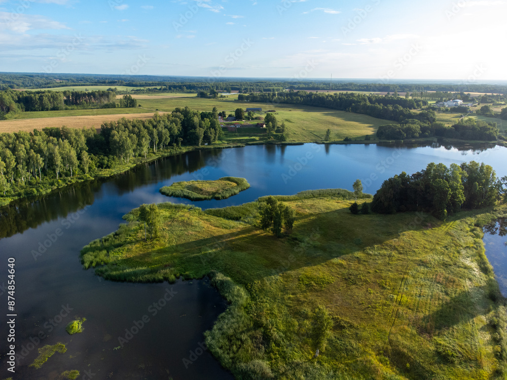 Fototapeta premium A beautiful summer landscape of a small lake, green trees and fields, taken from a drone. Natural rural landscape of Latvia, Northern Europe.
