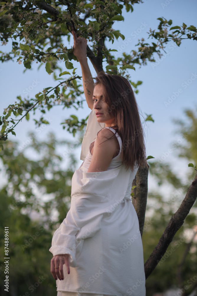 Naklejka premium A woman in a white outfit stretches her arms upwards, leaning against a tree. This image showcases the peaceful and harmonious interaction with the natural environment.
