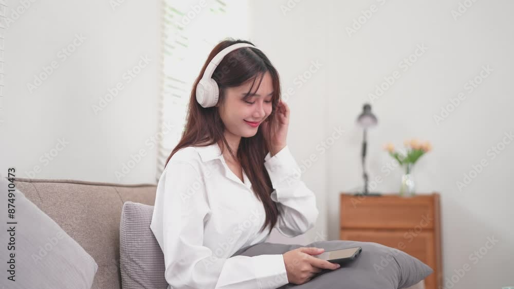 woman is sitting on a couch in a cozy living room, wearing white headphones and smiling as she holds a tablet. She is dressed in a white shirt and appears to be enjoying music or a podcast woman is sitting on a couch in a cozy living room, wearing white headphones and smiling as she holds a tablet. She is dressed in a white shirt and appears to be enjoying music or a podcast