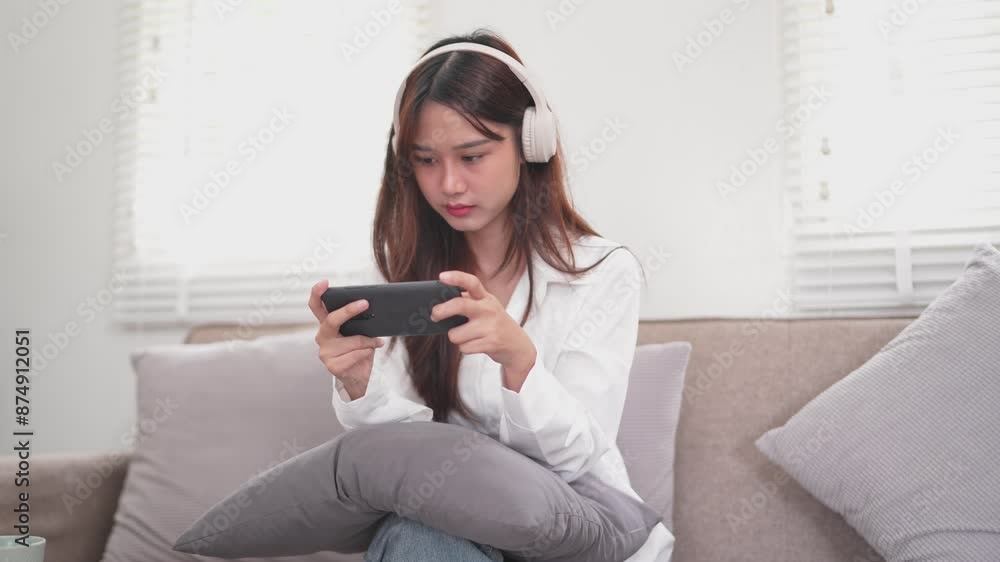 young woman is wearing white headphones and intently looking at her smartphone she is focused on watching a video or playing a game young woman is wearing white headphones and intently looking at her smartphone she is focused on watching a video or playing a game
