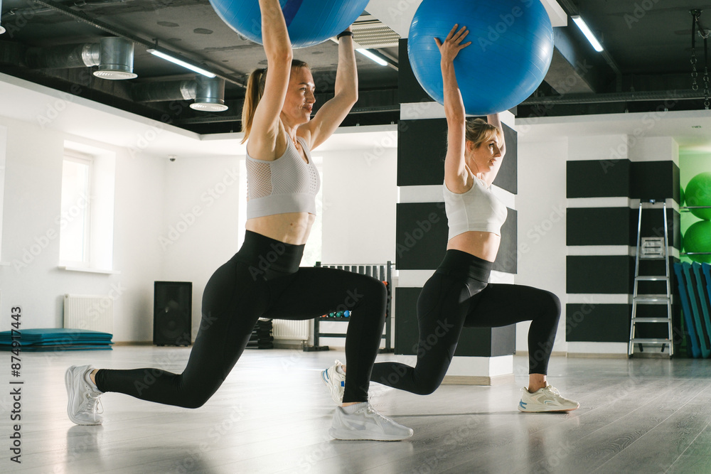 Obraz premium Two women are performing lunges with fitballs in a gym. Wearing athletic clothing and have their legs bent at a 90-degree angle. Focused on workout