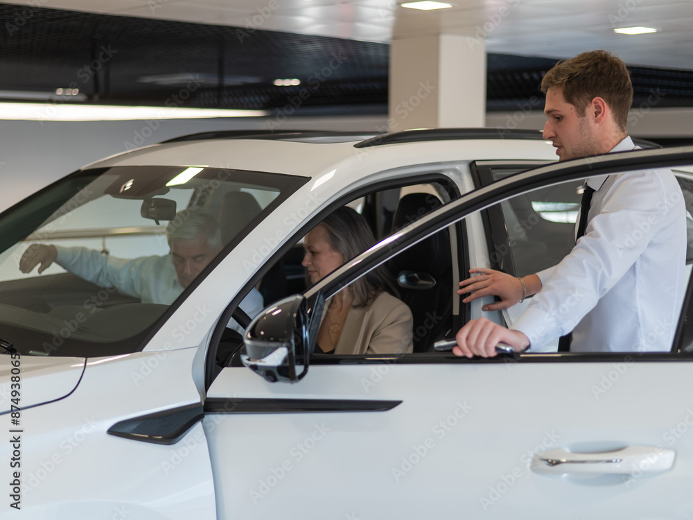 A gray-haired Caucasian woman is sitting behind the wheel of a car. Car dealership salesman consulting mature married couple. A gray-haired Caucasian woman is sitting behind the wheel of a car. Car dealership salesman consulting mature married couple.