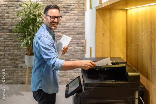 Portrait of male printer smiling and putting sheet of paper into photocopier machine in modern office