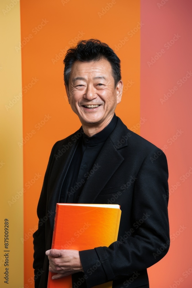 A smiling senior man in a black suit holds an orange book against a colorful background A smiling senior man in a black suit holds an orange book against a colorful background