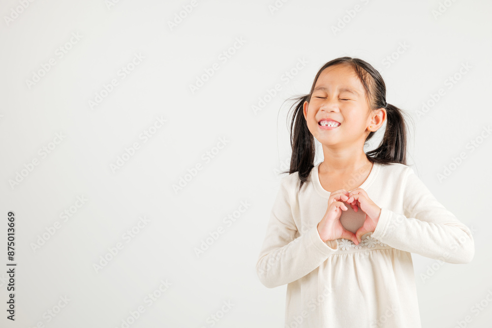 A smiling Asian young kid girl shows confidence, creating a heart shape with fingers and hands on white background. Portrait of kindergarten child sending love for Valentine's Day A smiling Asian young kid girl shows confidence, creating a heart shape with fingers and hands on white background. Portrait of kindergarten child sending love for Valentine's Day
