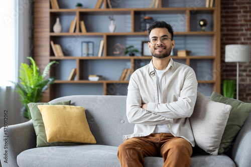 Smiling young Indian man sitting on sofa at home, arms crossed on chest and looking confidently at camera