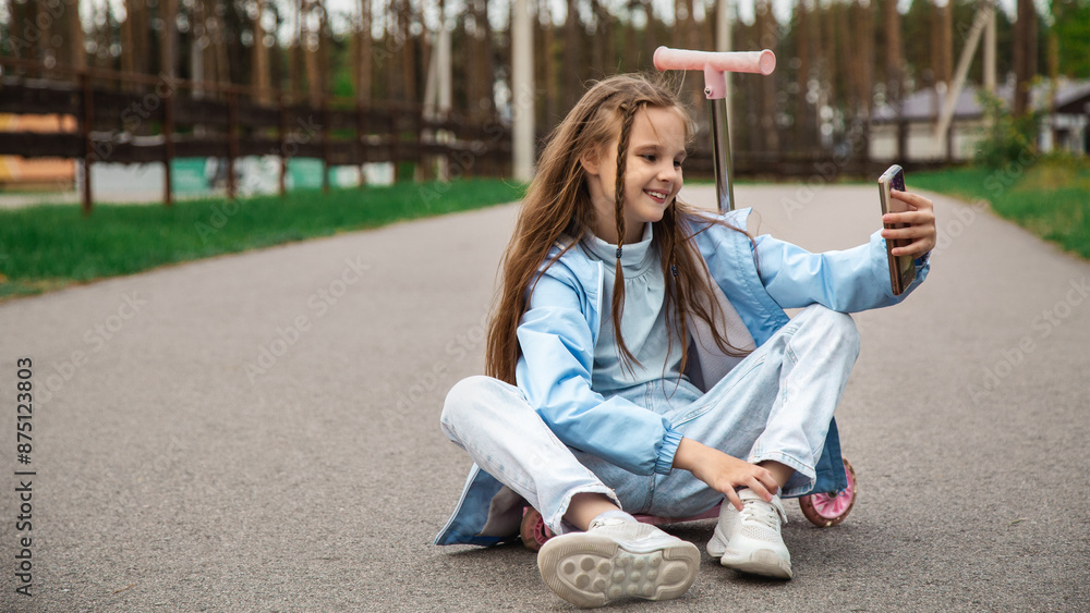 Beautiful little girl riding her scooter and taking a selfie on a mobile phone, in a park outdoors Beautiful little girl riding her scooter and taking a selfie on a mobile phone, in a park outdoors