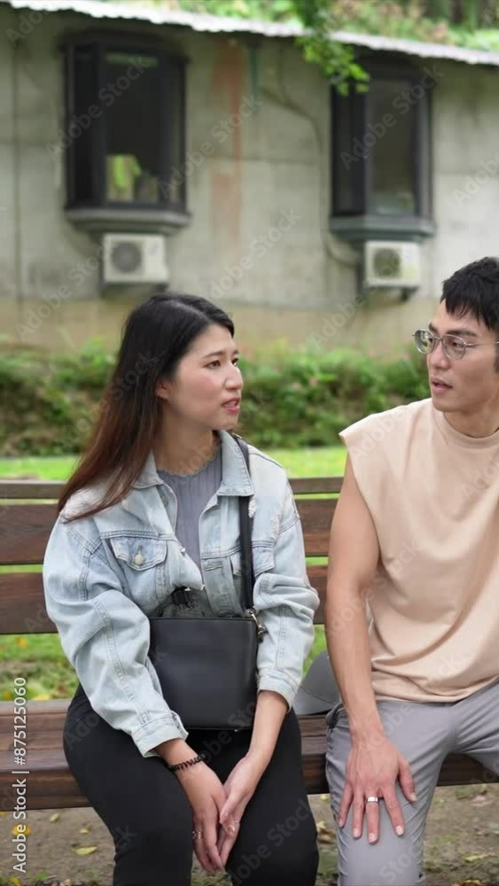 A vertical slow-motion video of a young Taiwanese couple in their twenties sitting on a bench and talking in a park in Xinyi District, Taipei City, on a spring afternoon.