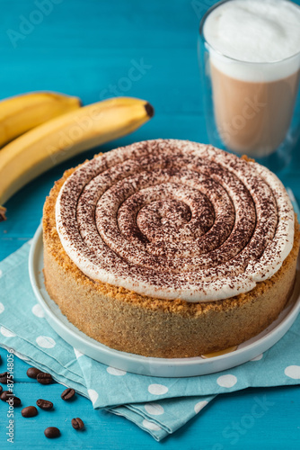 Banoffe pie filled with banana and dulce de leche and sprinkled with grated chocolate. Banana cake on white plate with glass of coffee on turquoise summer background photo