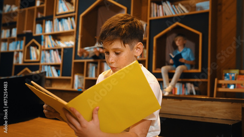 Caucasian boy reading a book while group of smart students sitting at library. Attractive child studying, learning from novel or textbook while children talking, chitchat about education. Erudition. photo