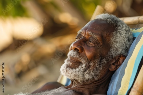 A senior man with white hair is seen leaning back on a beach chair, enjoying the tranquil setting of the beachfront, with background trees adding a touch of nature. photo