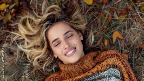Blonde woman dressed in a warm crocheted sweater, lying on a grassy field with her eyes closed, surrounded by autumn leaves, capturing a serene and happy moment in nature.