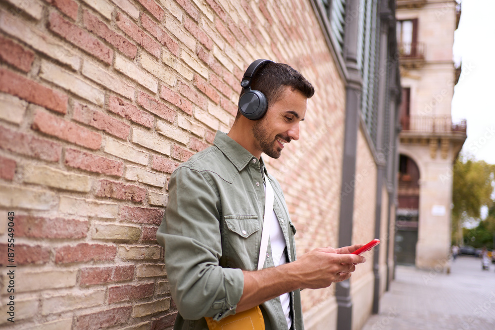 Obraz premium Attractive young Caucasian man with wireless headphones using cell phone leaning against brick wall. Millennial male looking happy at mobile while waiting for someone on city street. Copy space