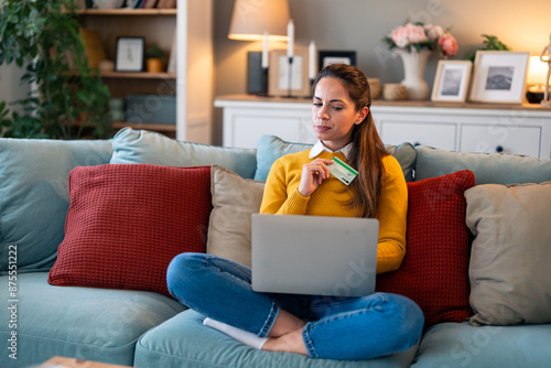 Cuadro en lienzo Young woman looking suspicious at laptop screen while holding credit card and sitting on a couch at home