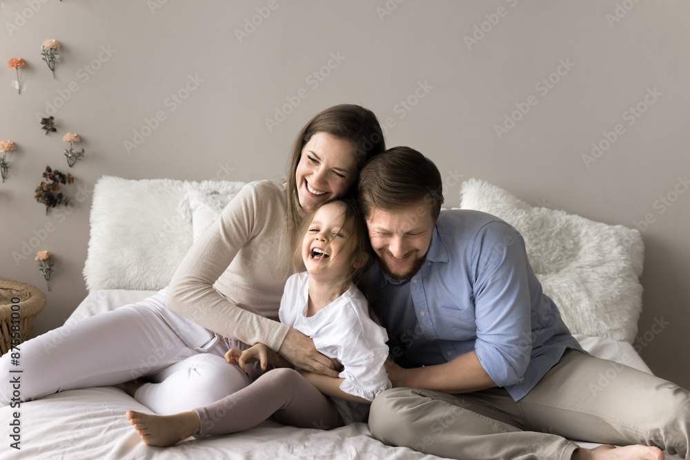 Young laughing parents spend time with preschooler daughter, sitting together on the bed, tickling adorable cheery child, enjoy funny active playtime at home. Family pastime, ties and happy parenthood Young laughing parents spend time with preschooler daughter, sitting together on the bed, tickling adorable cheery child, enjoy funny active playtime at home. Family pastime, ties and happy parenthood