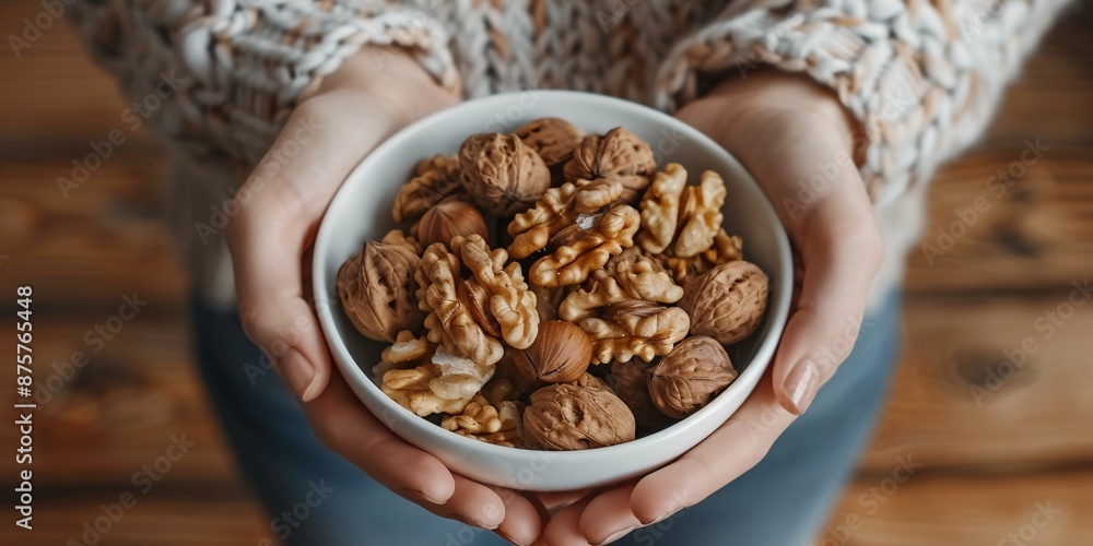 Obraz premium A dish of complete walnuts held by a woman, with a handful of unpeeled nuts in a white bowl on a wooden background, for nourishing and nutritious eating, captured in a top-down, high-resolution image.
