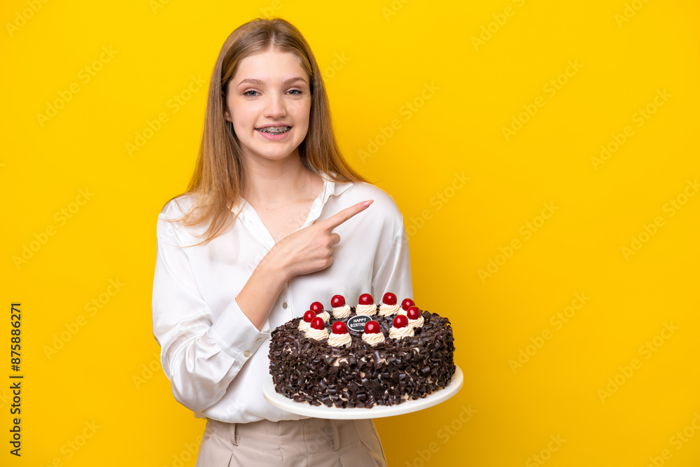Teenager Russian girl holding birthday cake isolated on yellow background pointing to the side to present a product Teenager Russian girl holding birthday cake isolated on yellow background pointing to the side to present a product