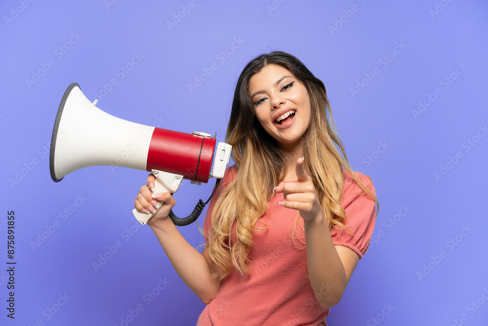 Young Russian girl isolated on blue background holding a megaphone and smiling while pointing to the front Young Russian girl isolated on blue background holding a megaphone and smiling while pointing to the front