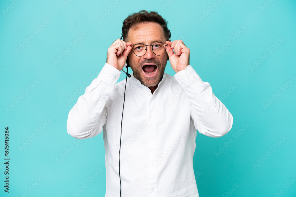 Telemarketer caucasian man working with a headset isolated on blue background with glasses and surprised Telemarketer caucasian man working with a headset isolated on blue background with glasses and surprised