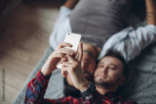 Portrait of two happy gay students smiling, taking selfies, lying on bed head to head in casual wear, having fun and posting photographs on social networks using smart phone. People and technology photo