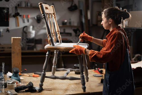 Young woman painting old wooden chair in grey color with paint brush working in her workshop. Restoration of the chair. Painting furniture. renovation, diy and furniture restoration