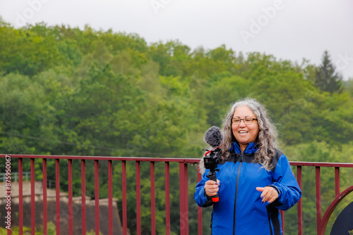 Smiling senior adult woman making videos with her camera, standing on dam of Robertville, green foliage of trees in misty and blurry background, blue jacket, cloudy day in Waimes, Belgium photo