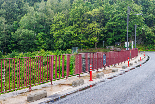 Empty rural road passing over Robertville Dam wall, leafy trees with green foliage in misty blurred background, cloudy day in Waimes, Belgium photo