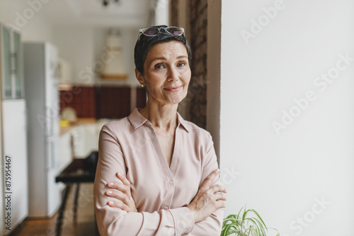 Charming pretty stylish real estate agent or home owner in elegant shirt with glasses on head smiling at camera standing with crossed hands in front of window, waiting for prospective clients