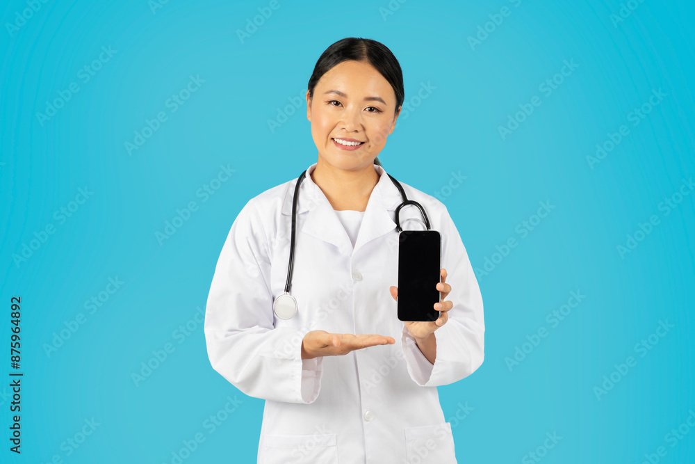 Asian woman doctor, wearing a white lab coat and stethoscope, stands against a blue background. She is smiling and holding a black smartphone with a blank screen in her right hand Asian woman doctor, wearing a white lab coat and stethoscope, stands against a blue background. She is smiling and holding a black smartphone with a blank screen in her right hand