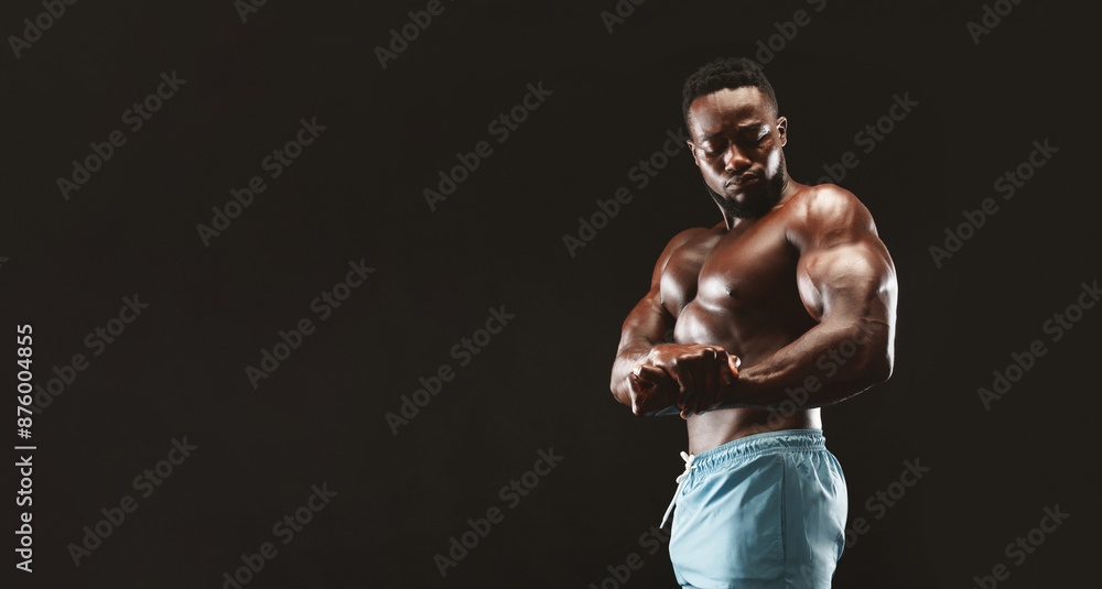 Naklejka premium African bodybuilder flexing his muscles, showing biceps in studio, black background