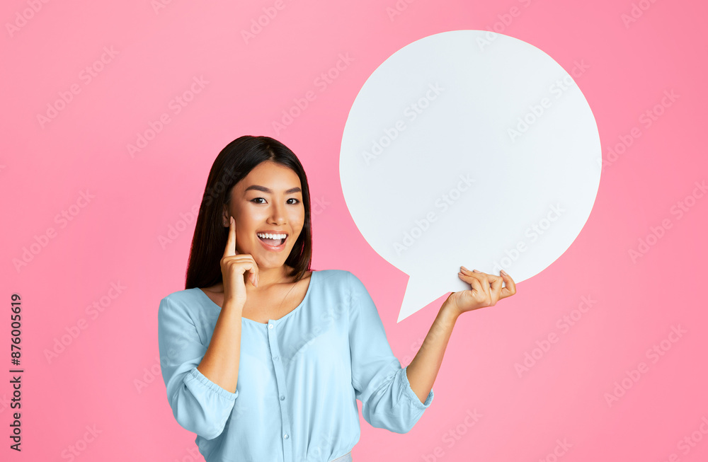 Asian smiling young woman standing in front of a pink background, holding a blank speech bubble in her right hand. Her left hand is touching her cheek Asian smiling young woman standing in front of a pink background, holding a blank speech bubble in her right hand. Her left hand is touching her cheek