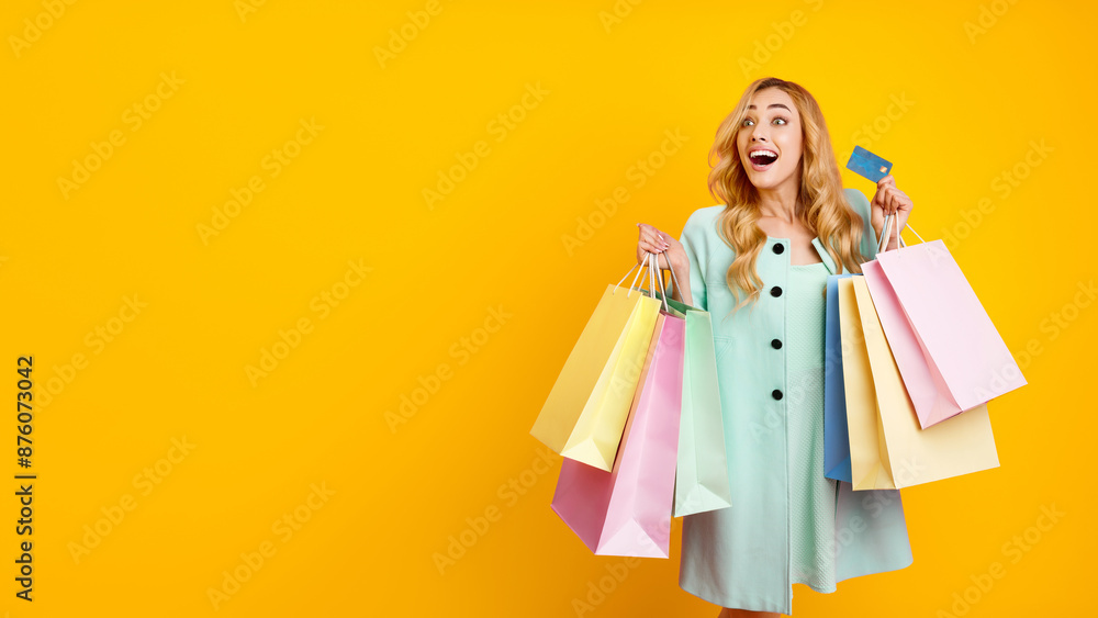 This image features a happy woman holding a stack of shopping bags and a credit card, against a bright yellow background. She is dressed in a light blue dress and has long blonde hair This image features a happy woman holding a stack of shopping bags and a credit card, against a bright yellow background. She is dressed in a light blue dress and has long blonde hair