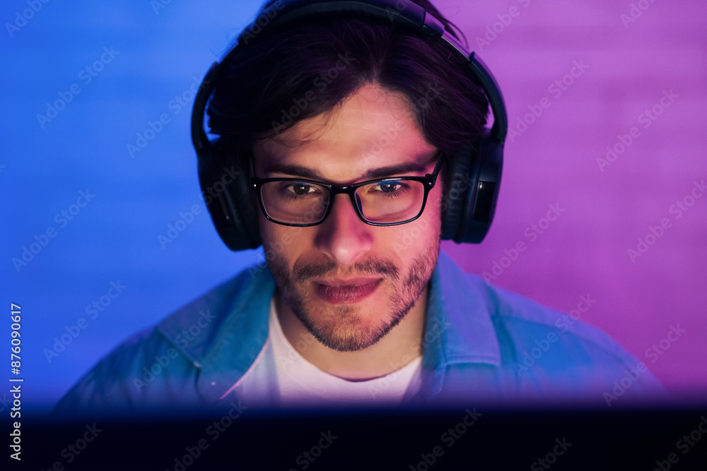 A young man is seen in close-up, wearing headphones and glasses, focused on a computer screen in front of him. The room is lit in blue and purple lighting A young man is seen in close-up, wearing headphones and glasses, focused on a computer screen in front of him. The room is lit in blue and purple lighting