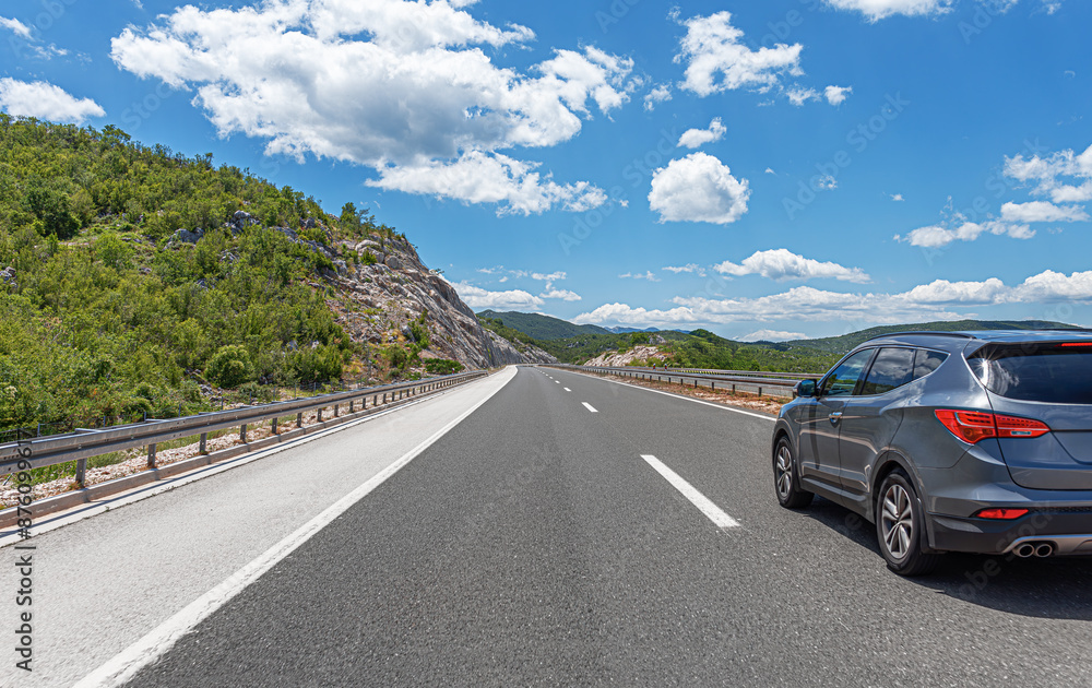 A grey car drives along the highway against the backdrop of rocky mountains on a sunny day.