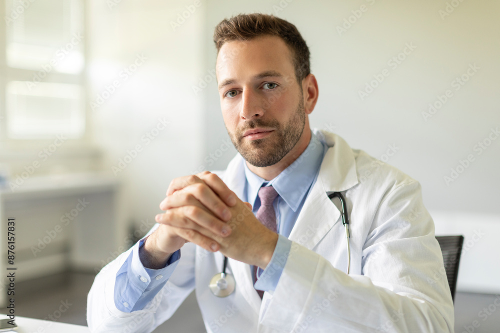 Serious professional man doctor in white uniform sitting at desk in clinic and looking at camera Serious professional man doctor in white uniform sitting at desk in clinic and looking at camera